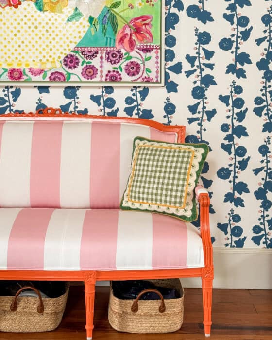 Orange painted settee with pink and white striped fabric in front of a blue hollyhock wallpaper.