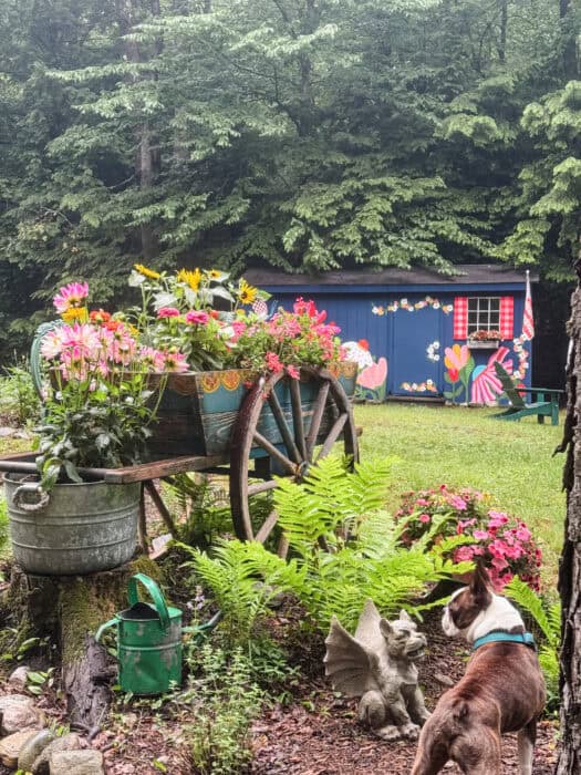 Antique wooden wagon with folk art painting used as a garden planter filled with dahlias, zinnias, and sunflowers in front of a blue cabin
