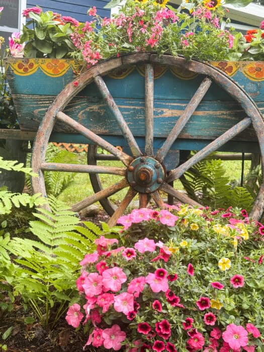 Antique wooden wagon with folk art painting used as a garden planter filled with dahlias, zinnias, and sunflowers in front of a blue cabin
