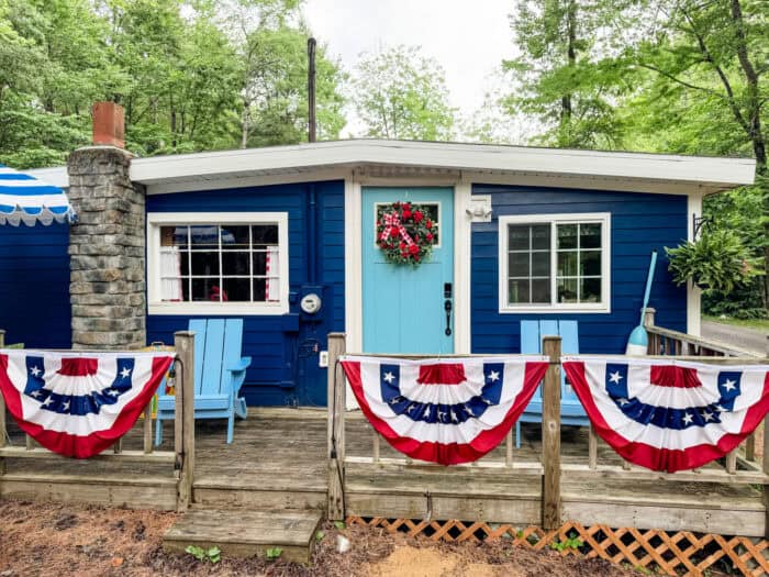 Cabin porch decorated for July 4th with red, white, and blue bunting and a patriotic wreath on a turquoise door.