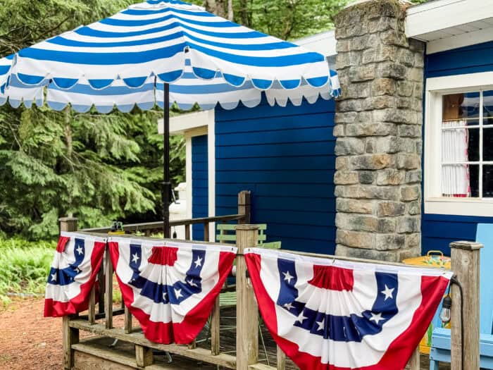 July 4th porch with American flag bunting, a striped umbrella, and a hanging fern holding a mini American flag.