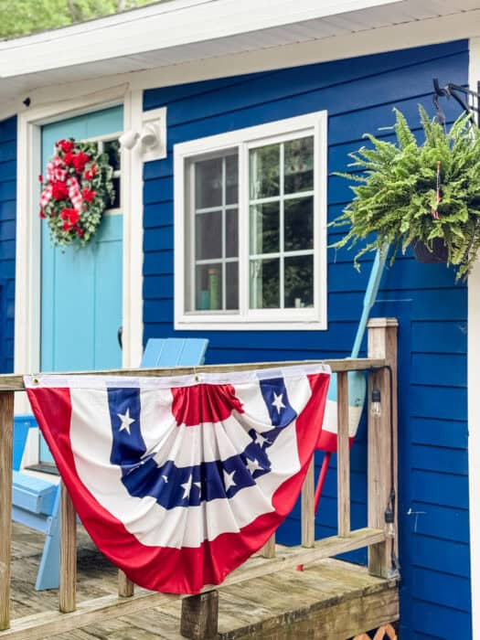 Red floral wreath with a gingham bow on a bright blue door, with patriotic bunting along a rustic wood porch railing.