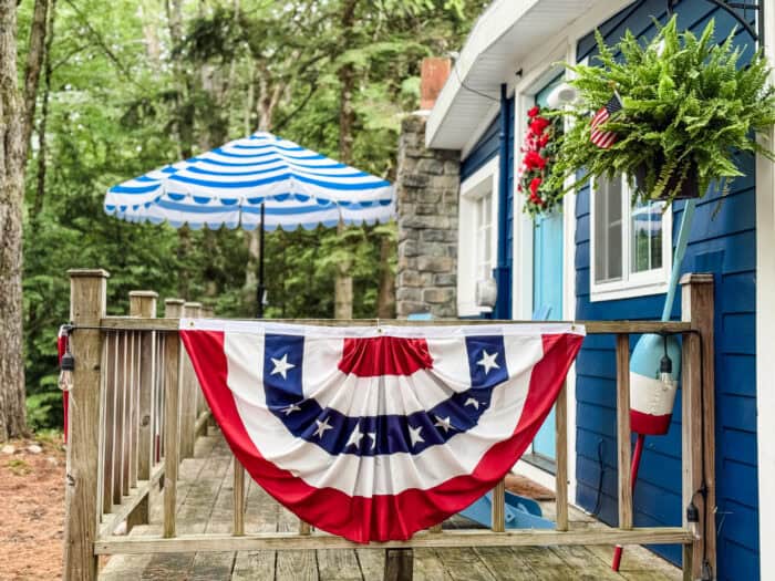 July 4th porch with American flag bunting, a striped umbrella, and a hanging fern holding a mini American flag.