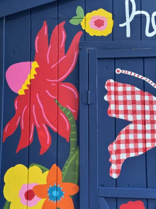 Navy shed covered in oversized, colorful flowers, butterflies, daisies, and gingham accents.