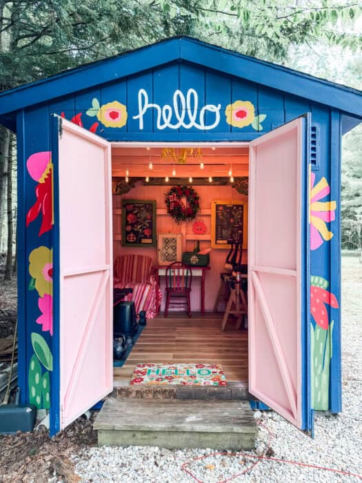 Pink shed interior makeover: blue shed with pink doors open, floral mural and “hello” sign.